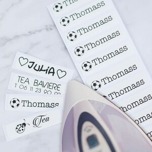 May include: White rectangular labels with black text and illustrations. Some labels feature the name "Thomass" with a soccer ball icon. Other labels have names like "Julia" and "Tea" with a ladybug illustration. An iron is partially visible.