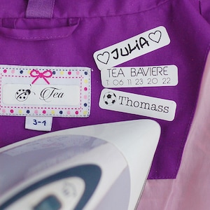 Peut inclure: Gros plan sur une veste violette avec plusieurs étiquettes thermocollantes. L'une porte le nom "Tea" avec une illustration de coccinelle et la taille "3-4". D'autres étiquettes affichent des noms et un ballon de foot. Un fer à repasser est au premier plan.