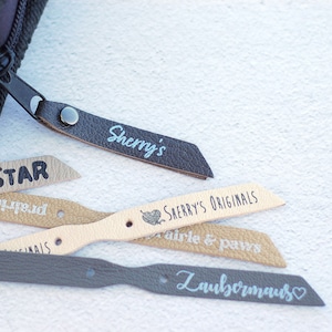 May include: A collection of leather zipper pulls with different names and phrases printed on them, including "Sherry's", "Sherry's Originals", "Up Star", "breathe &", "airle & paws", and "Zaubermaus".