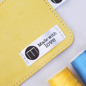 May include: A close-up shot of a yellow suede leather item with a stitched edge. A white tag with the words "Made with love" and a black circular logo is attached. Spools of colourful thread in yellow, blue, and purple are in the background.