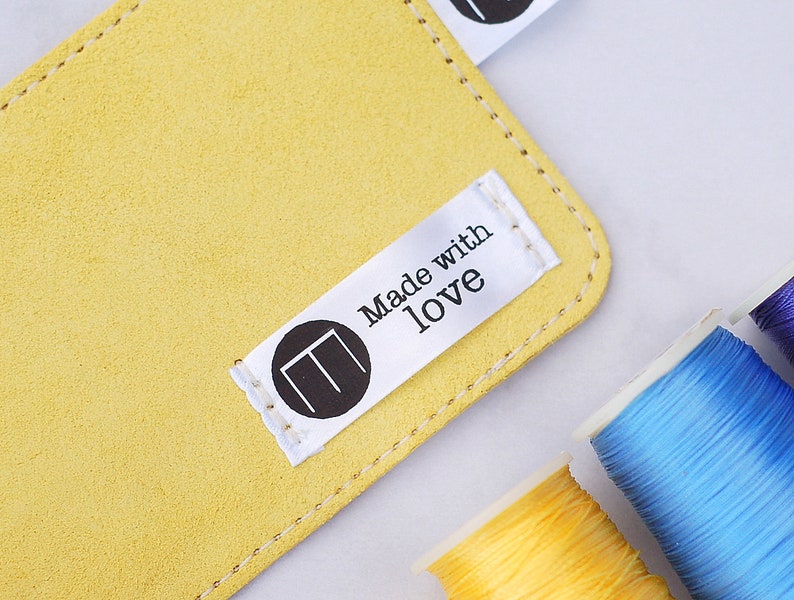 May include: A close-up shot of a yellow suede leather item with a stitched edge. A white tag with the words "Made with love" and a black circular logo is attached. Spools of colourful thread in yellow, blue, and purple are in the background.