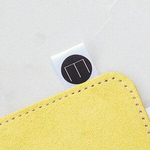 May include: Close-up of a yellow suede item with a stitched edge. A white label with a black circle and a white "M" design is attached to the item.