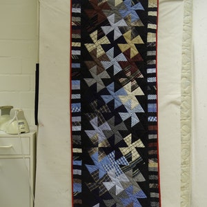 Könnte beinhalten: Eine lange, schmale Steppdecke mit schwarzem Hintergrund und rotem Rand. Die Steppdecke besteht aus Patchwork-Quadraten in Blautönen, Braun- und Weißtönen, die in einem Windmühlenmuster angeordnet sind.