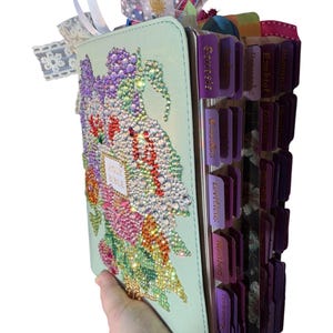 May include: A light green Holy Bible with a floral design made of rhinestones. The cover features a colorful arrangement of flowers and leaves. The book has purple tabs with book names and colorful ribbons at the top.