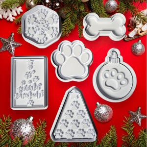 Könnte beinhalten: Satz mit sieben weißen Plastik-Ausstechformen in verschiedenen Formen, darunter ein Schneeflocke, ein Knochen, ein Pfotenabdruck, ein Weihnachtsbaum, ein Weihnachtsbaumschmuck und ein Baum mit Pfotenabdrücken. Die Ausstechformen befinden sich auf einem roten Hintergrund mit silbernen Ornamenten und Kiefernästen.