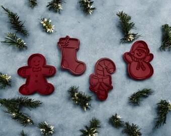 May include: Four red Christmas cookie cutters on a gray background. The cutters are shaped like a gingerbread man, a stocking, a candy cane, and a snowman.