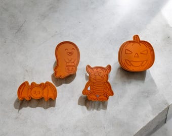 May include: Four orange plastic Halloween cookie cutters: a ghost, a bat, a monster, and a pumpkin.
