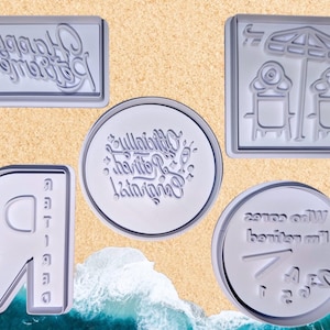 Op de afbeelding: Een verzameling heldere acryl stempels met verschillende ontwerpen. Eén stempel leest "Happy Birthday" in een cursief lettertype. Andere stempels tonen strandscènes en tekst. De stempels zijn geplaatst tegen een zanderige achtergrond.