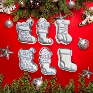 May include: Six white plastic Christmas stocking cookie cutters on a red background. The cutters feature different designs, including a stocking with candy canes, a stocking with a puppy, and a stocking with a snowman.