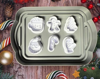 Puede incluir: Un juego de siete cortadores de galletas de Navidad de plástico blanco en una bandeja gris. Los cortadores tienen forma de hombre de jengibre, árbol de Navidad, bastón de caramelo, manopla, gorro de Papá Noel, taza de chocolate caliente y muñeco de nieve.