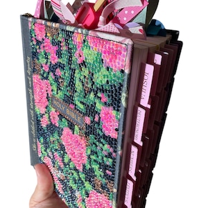 May include: A bedazzled Holy Bible with a floral design in pink, green, and black. The cover is embellished with sparkling rhinestones. Pink and green ribbons and tabs mark the pages. The text "HOLY BIBLE" is visible.