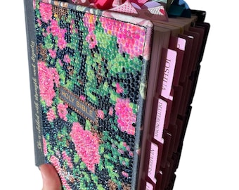 May include: A Holy Bible with a black, pink, and green floral rhinestone cover. The spine features the text She is clothed with strength and dignity. The book includes multiple colored ribbon bookmarks and indexed tabs labeled with book names like Joshua.