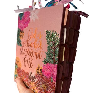May include: A pink Bible with floral embellishments and the text "God is within her she will not fall." The cover features colourful rhinestones and ribbon accents. The book has labelled tabs for easy navigation.