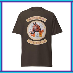 Puede incluir: Camiseta marrón con un diseño de cabeza de caballo dentro de una herradura, llamas y una estrella. El texto "KENDOM SALOON" está arqueado arriba y abajo de la ilustración del caballo. La camiseta es de color marrón liso.