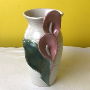 May include: A white ceramic vase with a green and pink floral design. The vase has two pink flowers with white centers and green leaves.