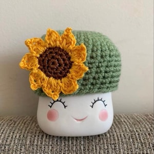 May include: A white ceramic mushroom-shaped planter with a smiling face, pink cheeks, and closed eyelashes. It has a green crocheted cap and a yellow and brown crocheted sunflower on top. The planter is sitting on a gray textured surface.
