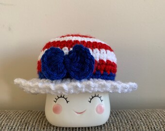 May include: A white ceramic mug with a smiling face, wearing a red, white, and blue striped crocheted hat with a blue bow.