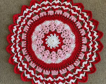 May include: A handmade, circular crochet doily in shades of red, white, and pink. The doily features a scalloped edge and concentric circles of varying patterns. The center is white, surrounded by pink and red, creating a decorative textile.