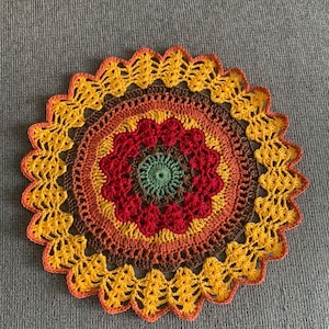 May include: A handmade, circular crochet doily in warm colors. The doily features concentric rings of red, orange, yellow, and brown, with a scalloped edge. The center is a small circle of green. This decorative item is perfect for home decor.