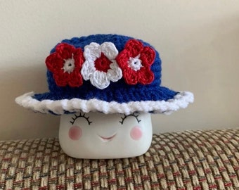 May include: A blue crocheted hat with white and red crocheted flowers. The hat is sitting on a white ceramic mug with a smiling face.