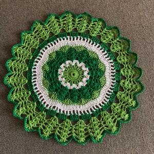 May include: A handmade, circular crochet doily in shades of green and white. The doily features a central floral design, surrounded by concentric rings of varying green hues and a white border. The outer edge has a scalloped, leaf-like pattern.