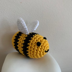 May include: A handmade crochet bee with yellow and black stripes. The bee has small black eyes and a smiling mouth. White wings are attached to the top of the bee's body. The bee is sitting on a white surface.