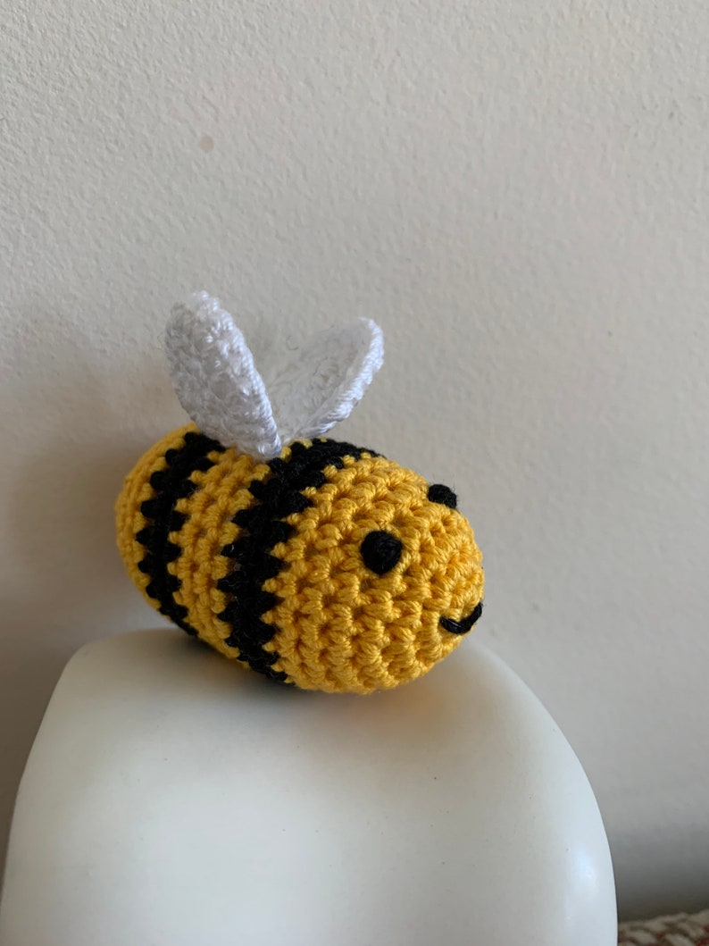 May include: A handmade crochet bee with yellow and black stripes. The bee has small black eyes and a smiling mouth. White wings are attached to the top of the bee's body. The bee is sitting on a white surface.