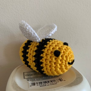 May include: A handmade crochet bee with yellow and black stripes, white wings, and small black eyes. The bee is a small, round, and whimsical decorative item. The bee is sitting on a white object.