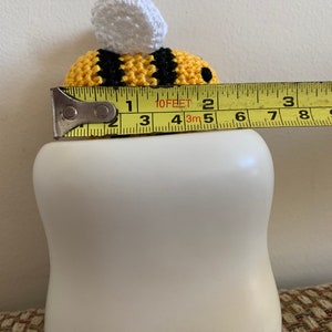 May include: A handmade crochet bee with yellow and black stripes and a white top, resting on a white ceramic object. The bee measures approximately 2 inches in length.