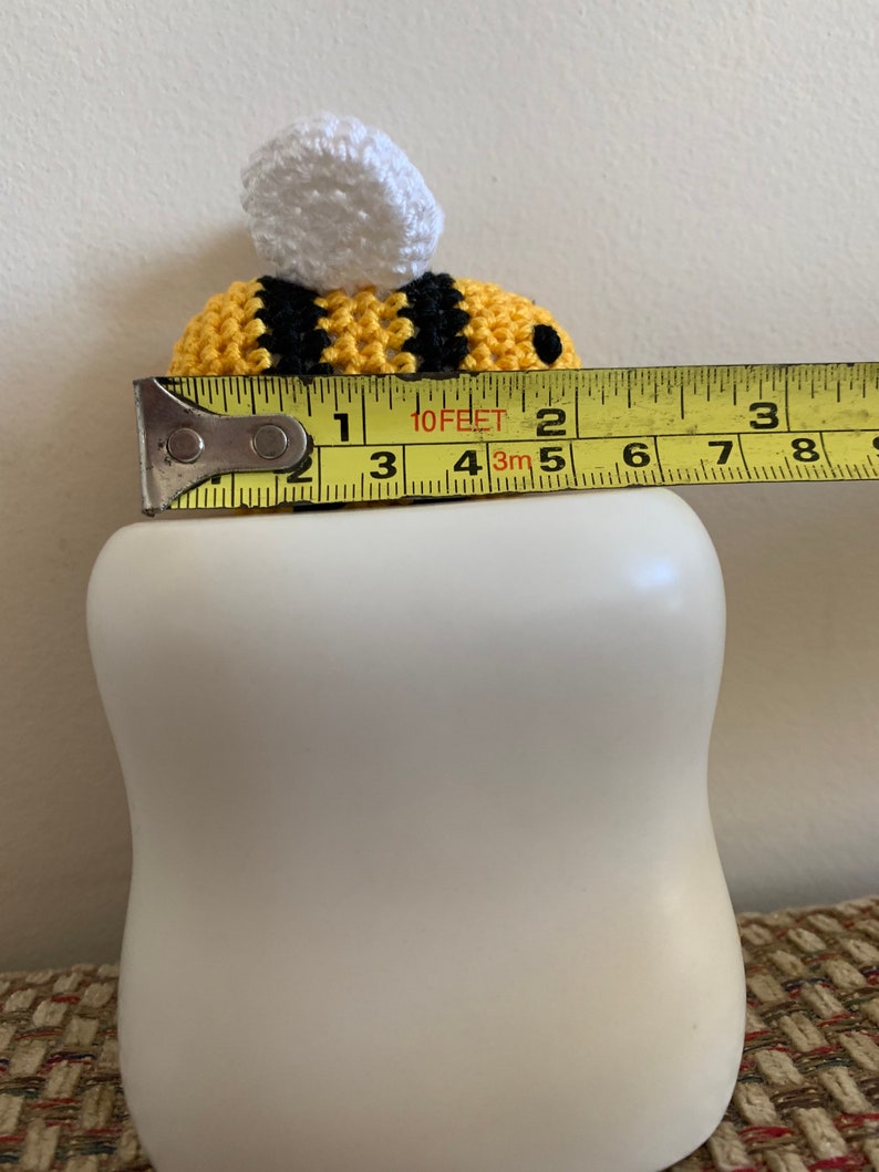 May include: A handmade crochet bee with yellow and black stripes and a white top, resting on a white ceramic object. The bee measures approximately 2 inches in length.