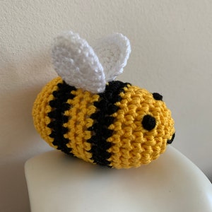 May include: A handmade crochet bee with yellow and black stripes. The bee has white wings and small black eyes. The bee is sitting on a white surface. The bee is a cute, whimsical item.