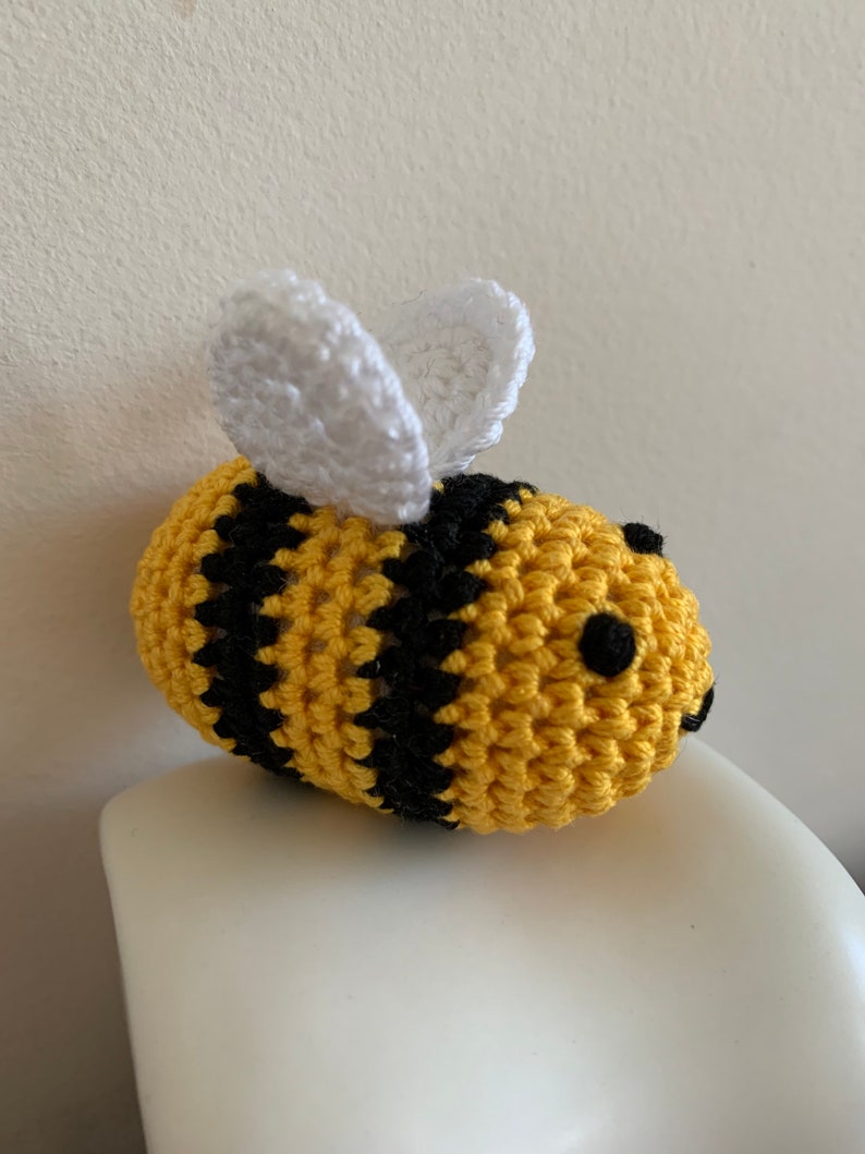 May include: A handmade crochet bee with yellow and black stripes. The bee has white wings and small black eyes. The bee is sitting on a white surface. The bee is a cute, whimsical item.