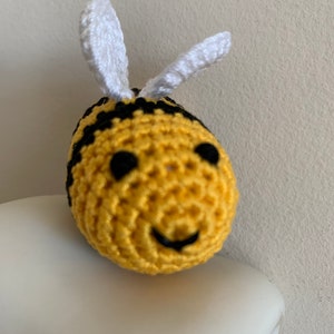 May include: A crocheted bee with a yellow body, black stripes, and white antennae. The bee has black eyes and a smiling mouth. The handmade item is a cute, whimsical decoration.