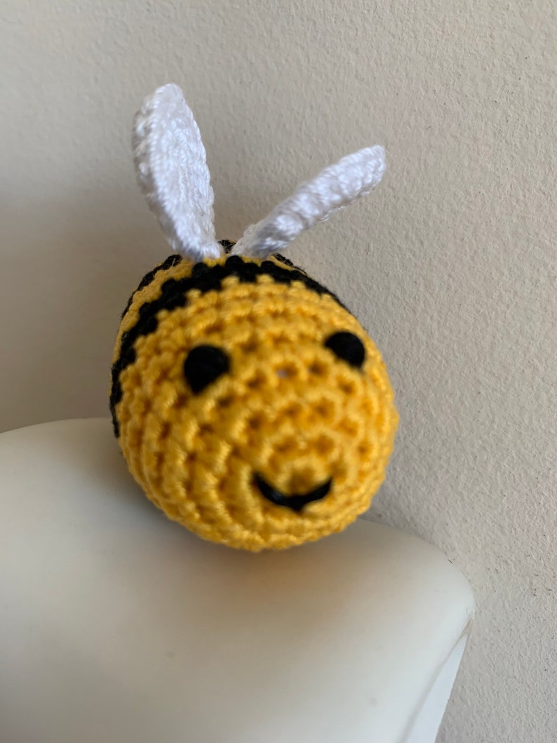 May include: A crocheted bee with a yellow body, black stripes, and white antennae. The bee has black eyes and a smiling mouth. The handmade item is a cute, whimsical decoration.