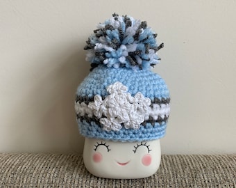 Könnte beinhalten: Eine hellblaue Häkelmütze mit einem weißen Bommel und einem weißen Häkelmuster. Die Mütze sitzt auf einer weißen Keramiktasse mit einem lächelnden Gesicht.