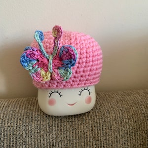 May include: A pink crocheted hat with a colorful butterfly design. The hat is sitting on a white ceramic mug with a smiling face.