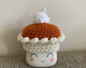 May include: A white ceramic mug shaped like a marshmallow with a crocheted pumpkin pie hat. The hat is brown with a white crocheted crust and a white whipped cream topping.