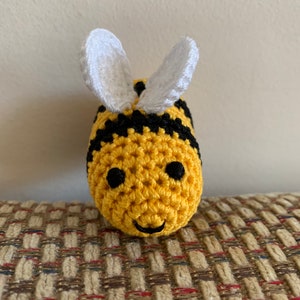 May include: A handmade crochet bee with a yellow body, black stripes, and white wings. The bee has black eyes and a smiling mouth. The bee is sitting on a patterned surface.