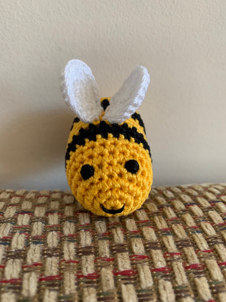 May include: A handmade crochet bee with a yellow body, black stripes, and white wings. The bee has black eyes and a smiling mouth. The bee is sitting on a patterned surface.