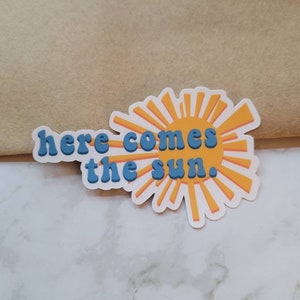 Puede incluir: Una pegatina con un sol amarillo y texto azul que dice "here comes the sun."