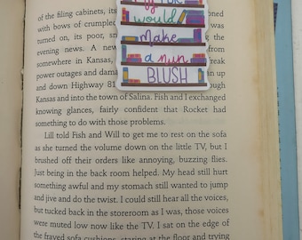 Op de afbeelding: Een kleurrijk bladwijzer met de tekst "My TBR would make a nun blush" en een boekenkastontwerp met boeken. De bladwijzer bevindt zich op een pagina van een boek.