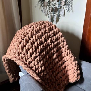 Könnte beinhalten: Eine grob gestrickte Decke in einem warmen, erdfarbenen Braunton. Die Decke ist über einen grauen Stuhl drapiert und zeigt ihr dickes, strukturiertes Gewebe. Die kuschelige Wohndecke ist perfekt, um jedem Wohnraum Wärme und Stil zu verleihen.