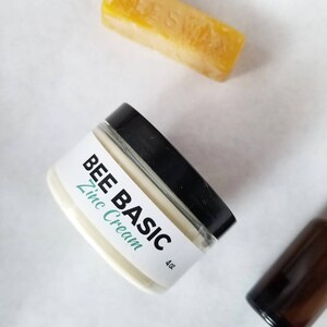 May include: A jar of "BEE BASIC Zinc Cream" with a black lid and a white label. The label includes the text "BEE BASIC" in black and "Zinc Cream" in a teal color. A yellow beeswax bar is in the background. The jar is 4 oz.