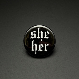 Puede incluir: Un botón redondo con alfiler que presenta una superficie negra brillante con la palabra "she" en la línea superior y "her" en la línea inferior, ambas impresas en una fuente blanca de estilo gótico centrada en la cara del botón.