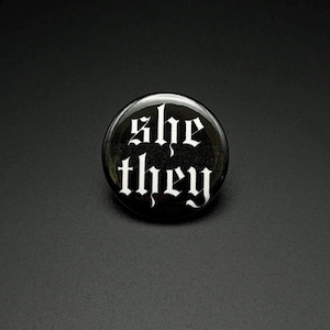 Puede incluir: Un botón circular con alfiler que presenta un fondo negro con el texto blanco "she" y "they" escrito en una fuente gótica estilizada. El botón tiene un acabado brillante y está centrado sobre un fondo oscuro y neutro.