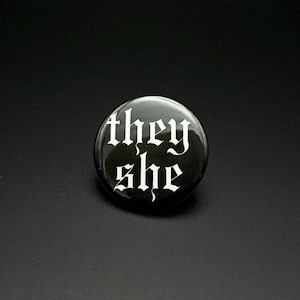 Puede incluir: Un botón circular con un fondo negro brillante y el texto blanco "they" en la línea superior y "she" en la línea inferior, ambos escritos en una fuente decorativa de estilo gótico con letras afiladas y ornamentadas.