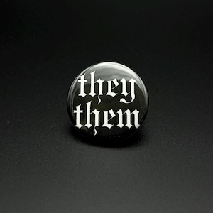 Puede incluir: Una chapa redonda con fondo negro y las palabras "they them" impresas en blanco con una tipografía gótica estilizada. La chapa tiene un acabado brillante que refleja la luz, creando un sutil punto de luz sobre su superficie.