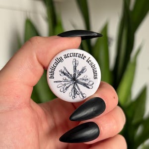 May include: A round white pin-back button featuring a black ink illustration of a stylised, multi-limbed figure at the centre. The text "biblically accurate lesbians" is printed in a black gothic-style font along the curved upper edge of the button.