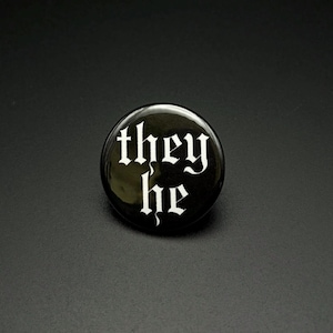 Puede incluir: Una chapa redonda con acabado negro brillante. El texto "they he" está impreso en el centro con una fuente gótica blanca estilizada. El diseño presenta un aspecto limpio y de alto contraste con letras blancas sobre el fondo oscuro.