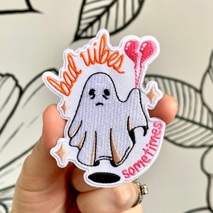 Puede incluir: Parche blanco bordado para planchar con un fantasma que sostiene un globo de corazón rosa. El texto dice "bad vibes sometimes" en letras naranja y rosa.